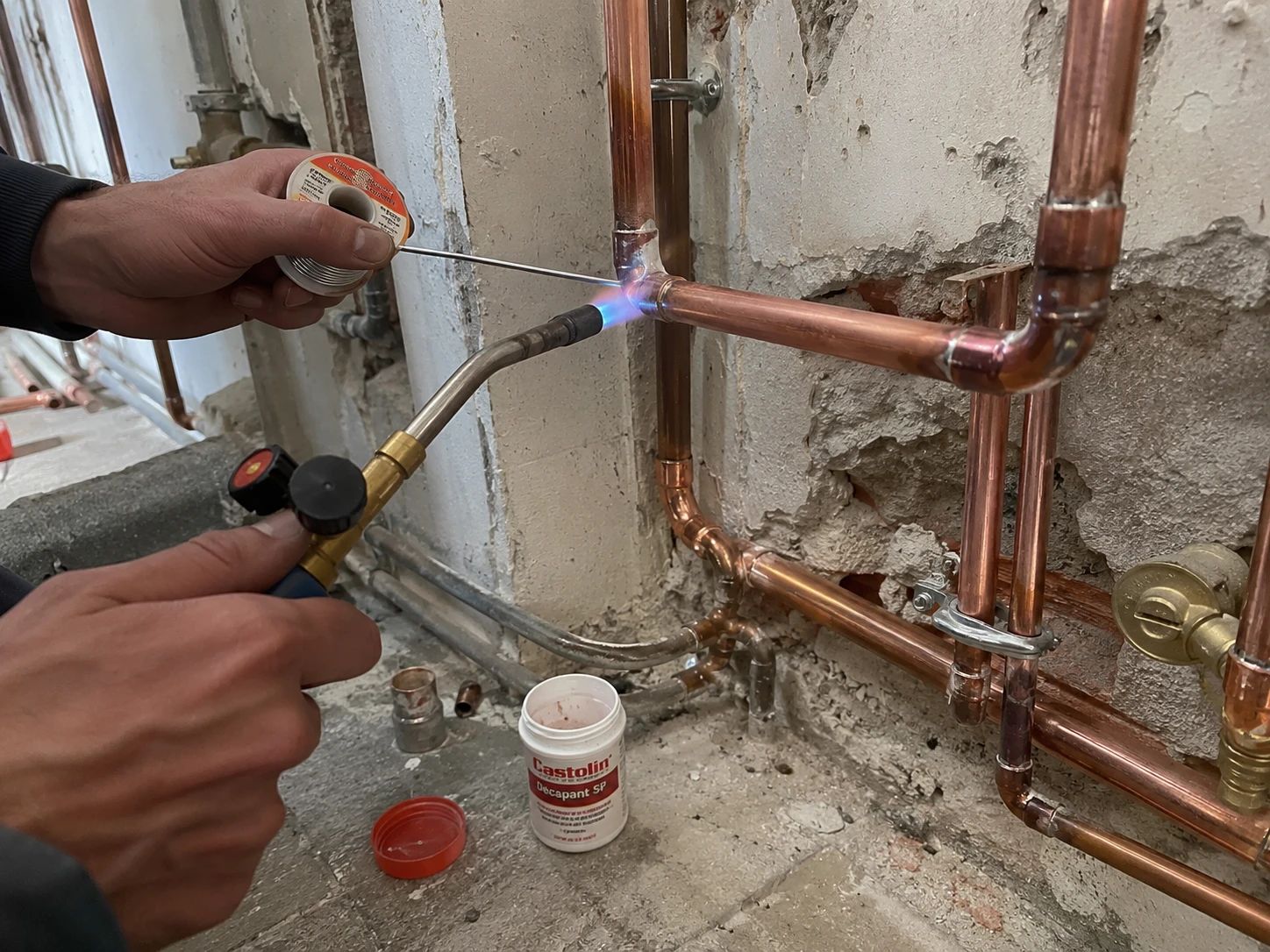 Plombier brasant un raccord en cuivre au chalumeau, décapant et fil de brasure, réseau fixé au mur par colliers