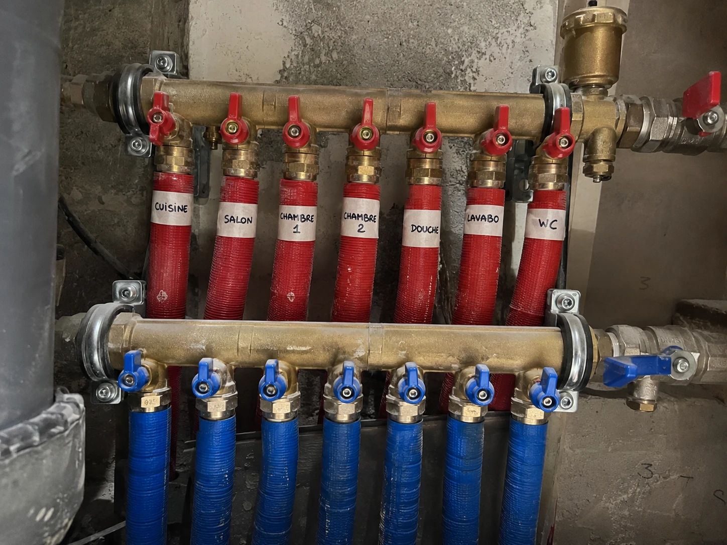 Collecteur en laiton avec vannes, départs eau chaude et eau froide étiquetés par pièce : cuisine, salon, chambres, douche, lavabo, WC