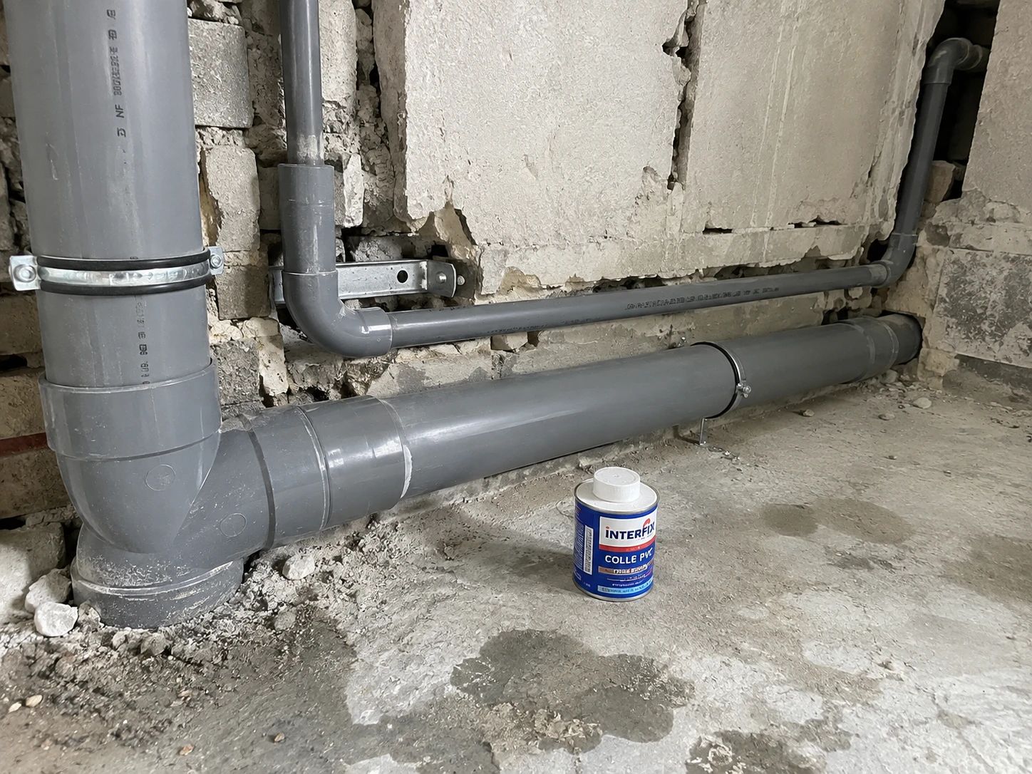 Évacuations en PVC gris Ø100 et Ø40 posées en pied de mur avec colliers, pot de colle PVC au sol